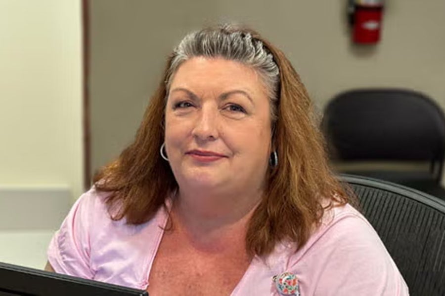 A woman wearing a pink shirt sits behind a computer while smiling up at the camera.