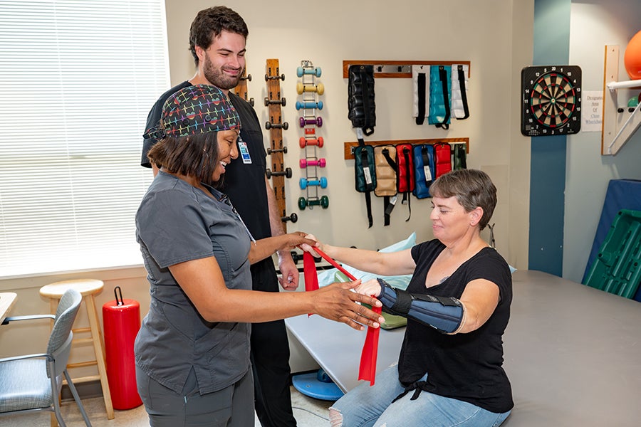 Two rehabilitation employees assist a patient who is sitting on a therapy bed stretching a resistance band with both of her arms.