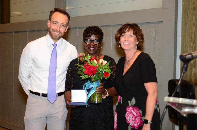 Cheryl Love, holding an award box and a vase of roses, stands between Russell Baxley and Colleen Duerr at the 2026 Employee Service Awards.