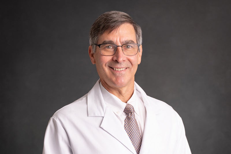 Professional headshot of Dr. Paul A. Slota wearing a white dress shirt, light colored, patterned tie and a white lab coat in front of a dark backdrop.