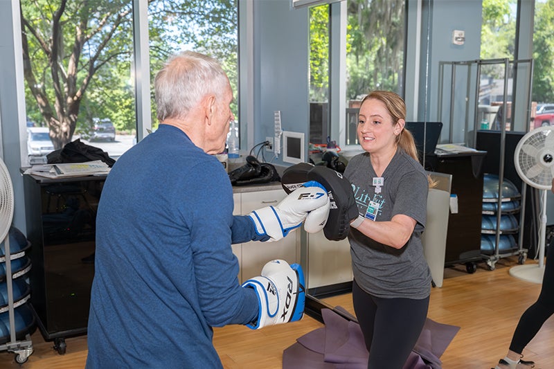 A Rock Steady Boxing instructor at LifeFit Wellness Center assists an older man with a boxing exercise.