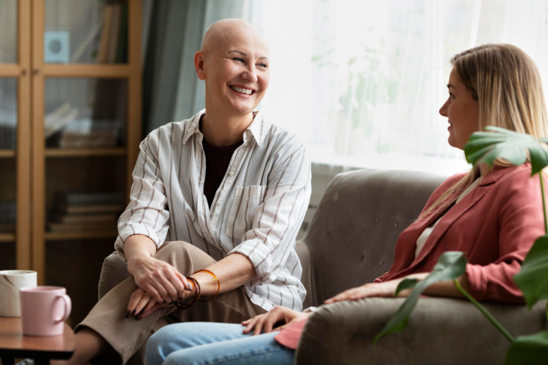 Female cancer patient with shaved head sits on a couch smiling at another woman on the other end of the couch.