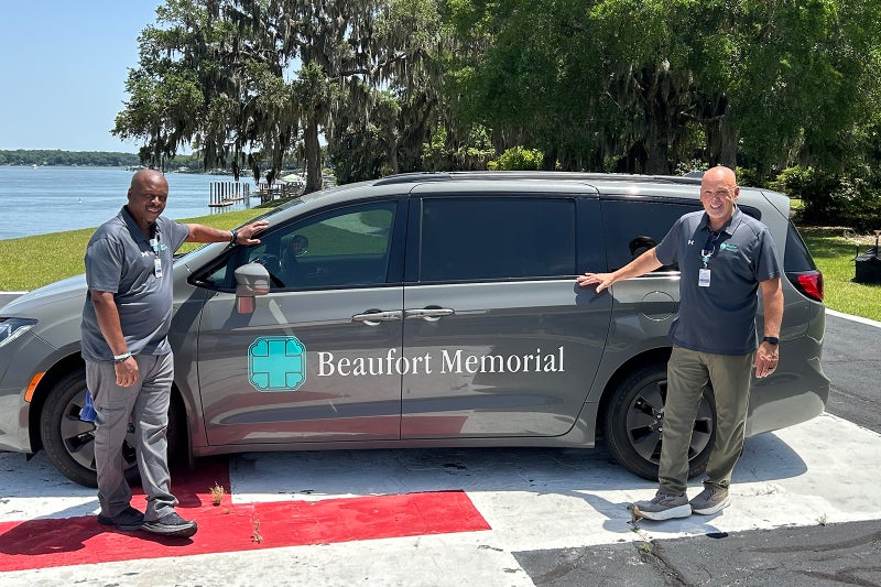 Two Beaufort Memorial transport department employees stand next to a gray minivan with the Beaufort Memorial logo on the side and a Lowcountry backdrop behind them