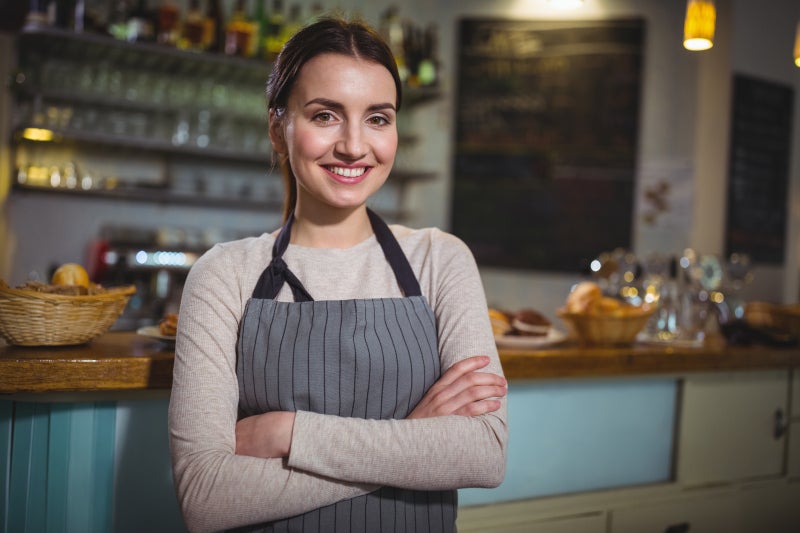 Young woman wearing an apron standing in front of restaurant counter with her arms crossed and smiling