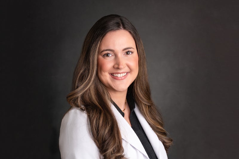 Lauren Nates, nurse practitioner, wears a black shirt with a white lab coat over in front of a darker grey backdrop.