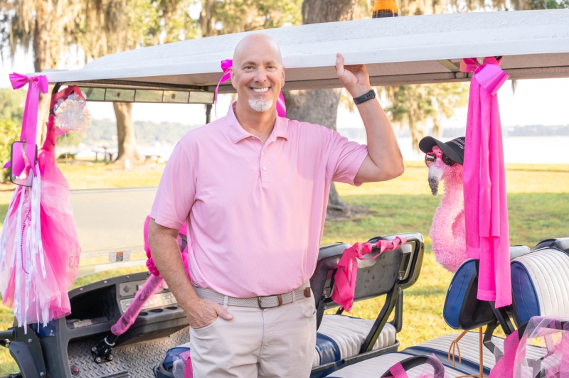 Dr. Eric Turner, an oncology surgeon with Beaufort Memorial Oncology Specialists, poses in pink casual attire next to a golf cart donned in pink decor with an outdoor backdrop.