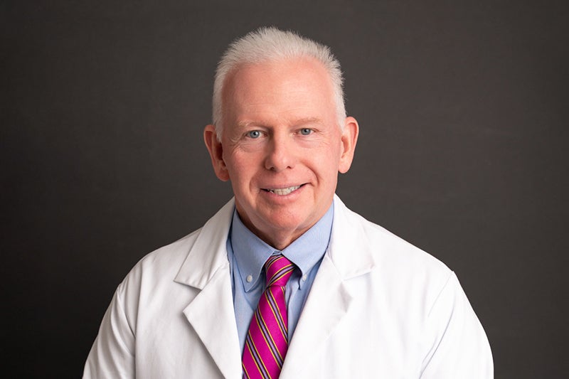 Dr. Glenn Mieszkalski poses in front of a dark grey backdrop wearing a dress shirt and tie with a white lab coat.