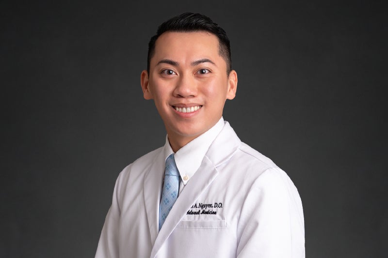 Smiling Dr. Minh Nguyen in front of a dark backdrop wearing his white lab coat embroidered with his name on the front left chest panel.