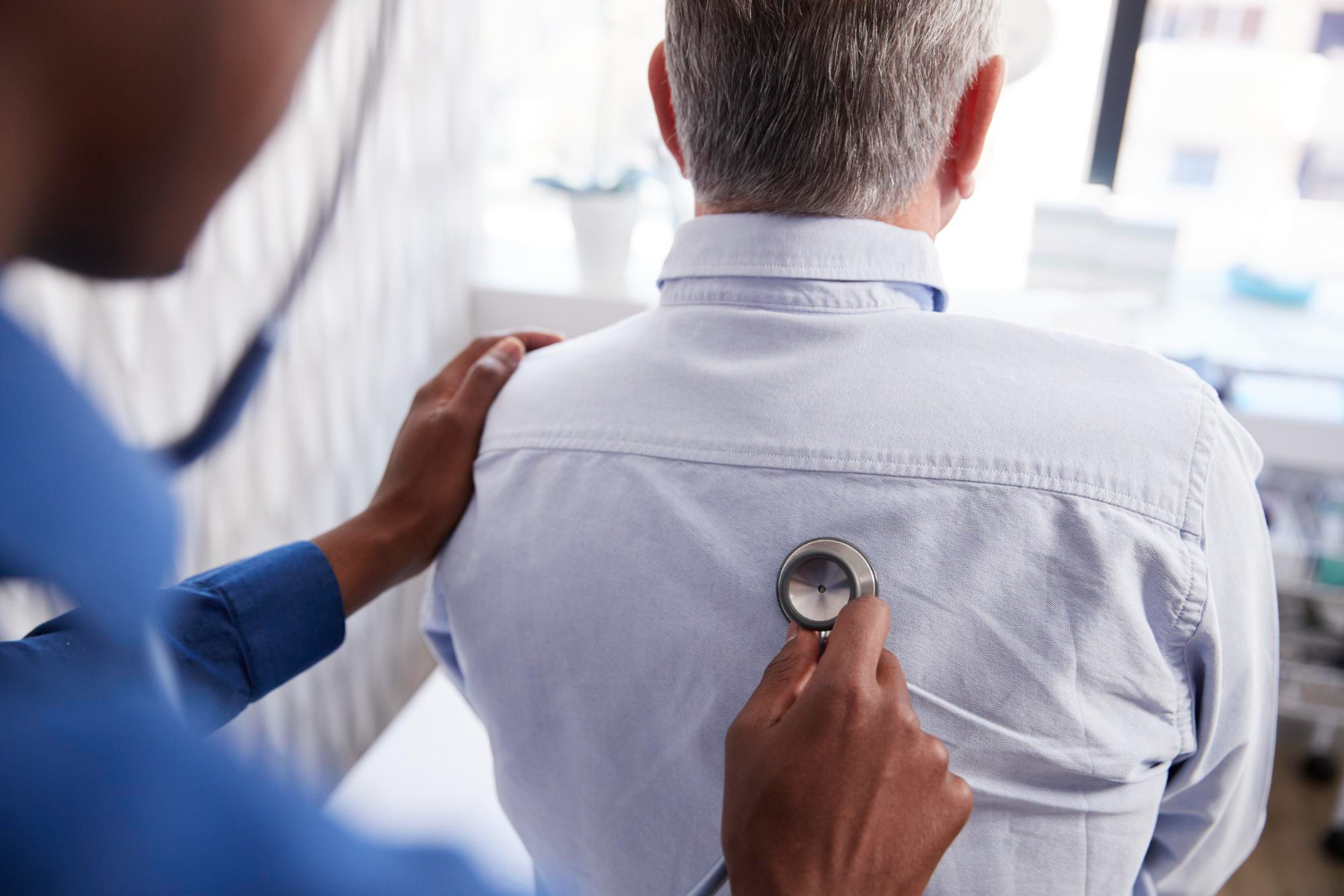 A medical expert listens to a male patient's lungs through a stethoscope positioned on the back