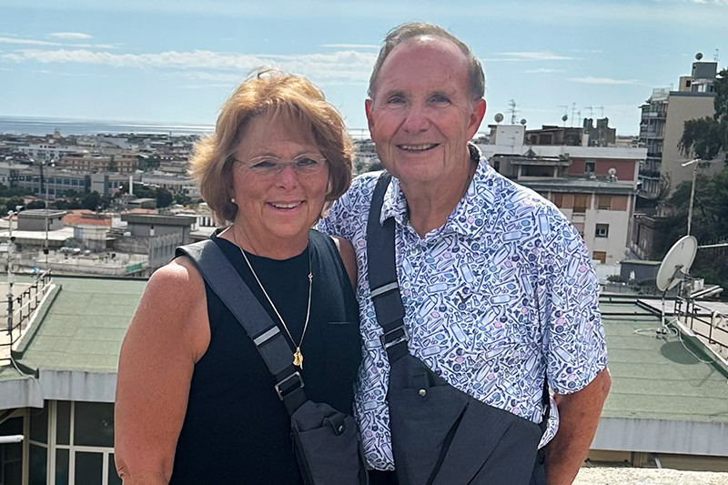 Grace Sorrentino and her husband pose for a photo with a Meditteranean country cityscape in the background