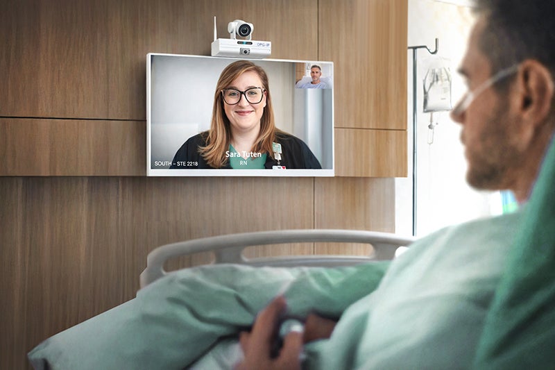 A middle aged man in a hospital bed looks out at a television screen that has a video call with Sarah Tuten, a registered nurse.