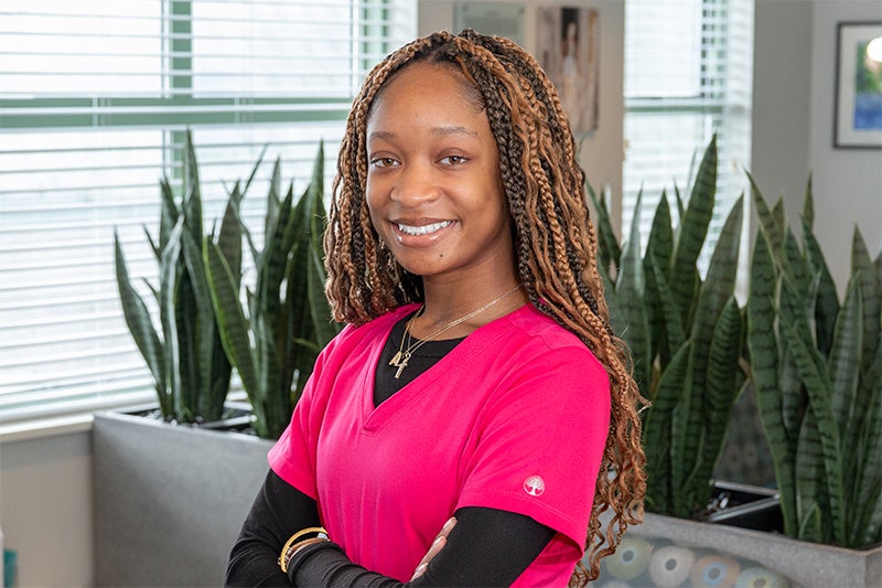 Alani Crawford, CPCT, poses in a healthcare clinic waiting room, wearing bold pink scrubs and a black long-sleeved shirt underneath.