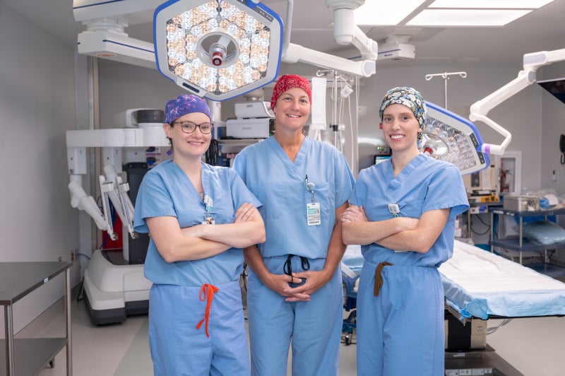 Drs. Taylor Turnbull, Deanna Mansker and Aubrey Place stand in a Beaufort Memorial operating room, all wearing light blue scrubs, each donning their own colorful scrub cap.