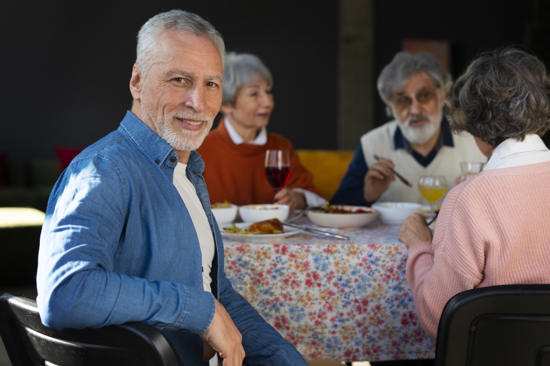An older male seated at a table with others turns to smile at the camera
