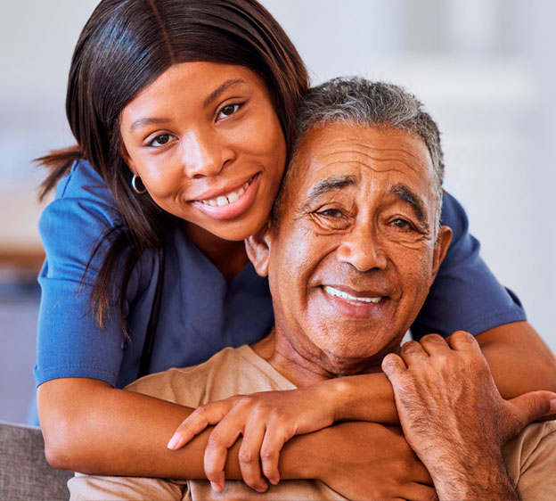 A young woman stands behind an older man, hugging him. They’re both smiling and looking into the camera.