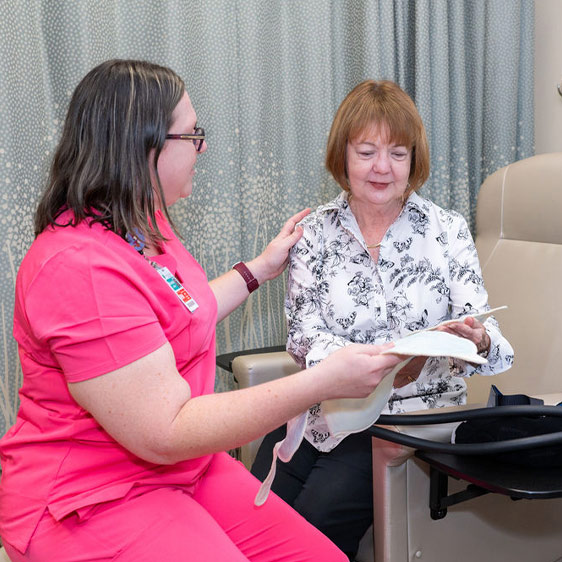 A woman named Terry Murray smiling at the camera and sitting, talking with another woman named Brandie Gillian, RN, while looking down at DigniCap