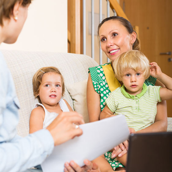 A woman sits on couch talking with a mom holding child