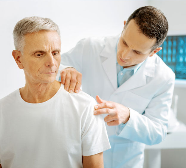 A male physician standing in front of a computer screen examines a male patient’s shoulder.
