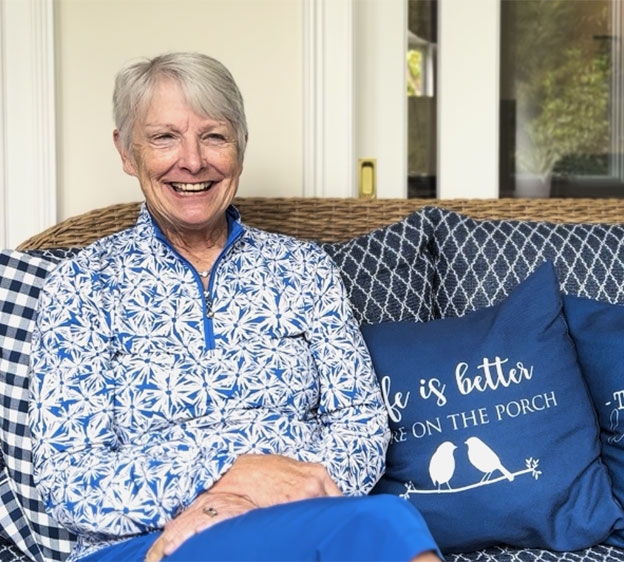 A woman named Sue Zankowski sitting on a wicker loveseat with blue cushions and smiling toward the camera.
