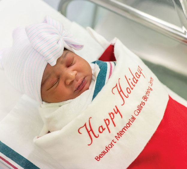 A photograph of a newborn wrapped in a stocking that says Happy Holidays, Beaufort Memorial Collins Birthing Center