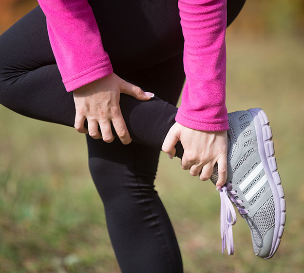 A woman wearing black running tights, a hot pink jacket and gray running shoes grabs her ankle.
