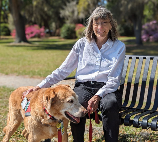 Beaufort Memorial volunteer Sunday Brown sits on a park bench while her therapy dog, Bella, stands next to her. Sunday smiles at the camera.