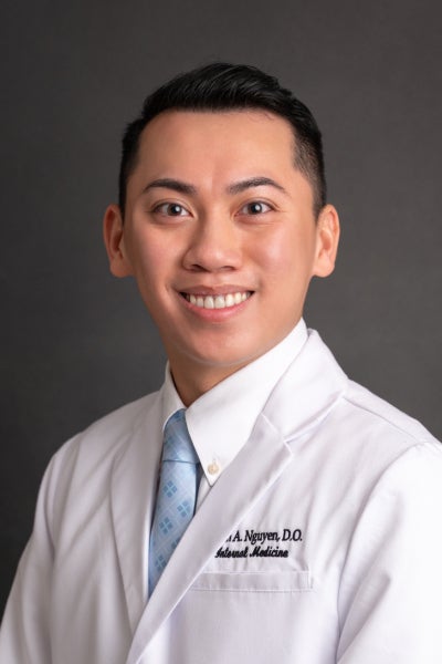 Dr. Minh Nguyen wears a white lab coat posing in front of a dark grey backdrop.